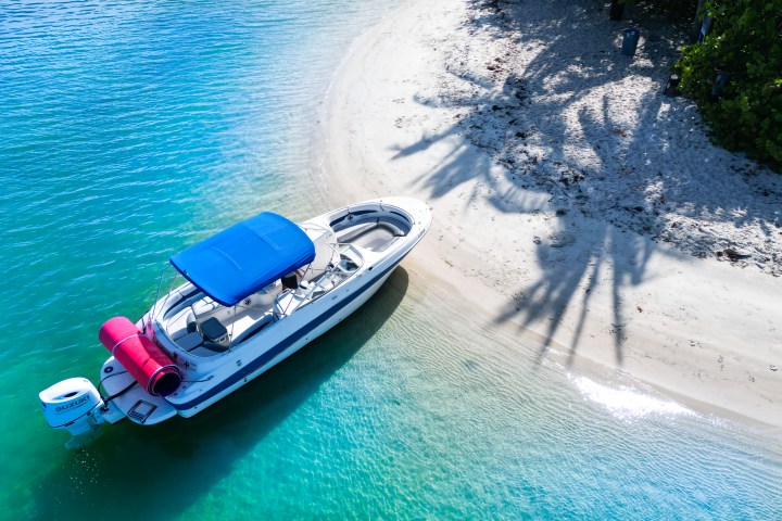 Motorboat with blue canopy near sandy beach and palm tree shadows on clear water.